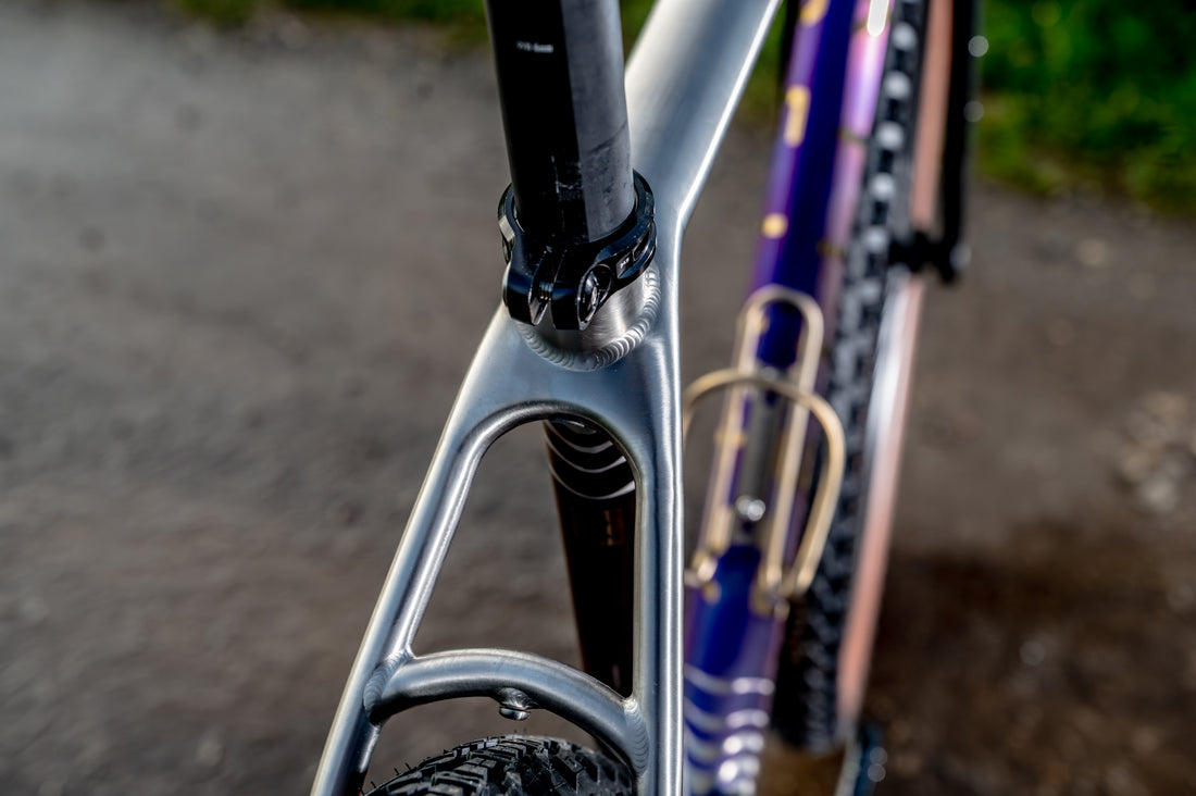 Close-up of a bicycle frame showing a silver metallic down tube and seat tube junction, with a purple and black rear wheel visible in the background on an outdoor path.