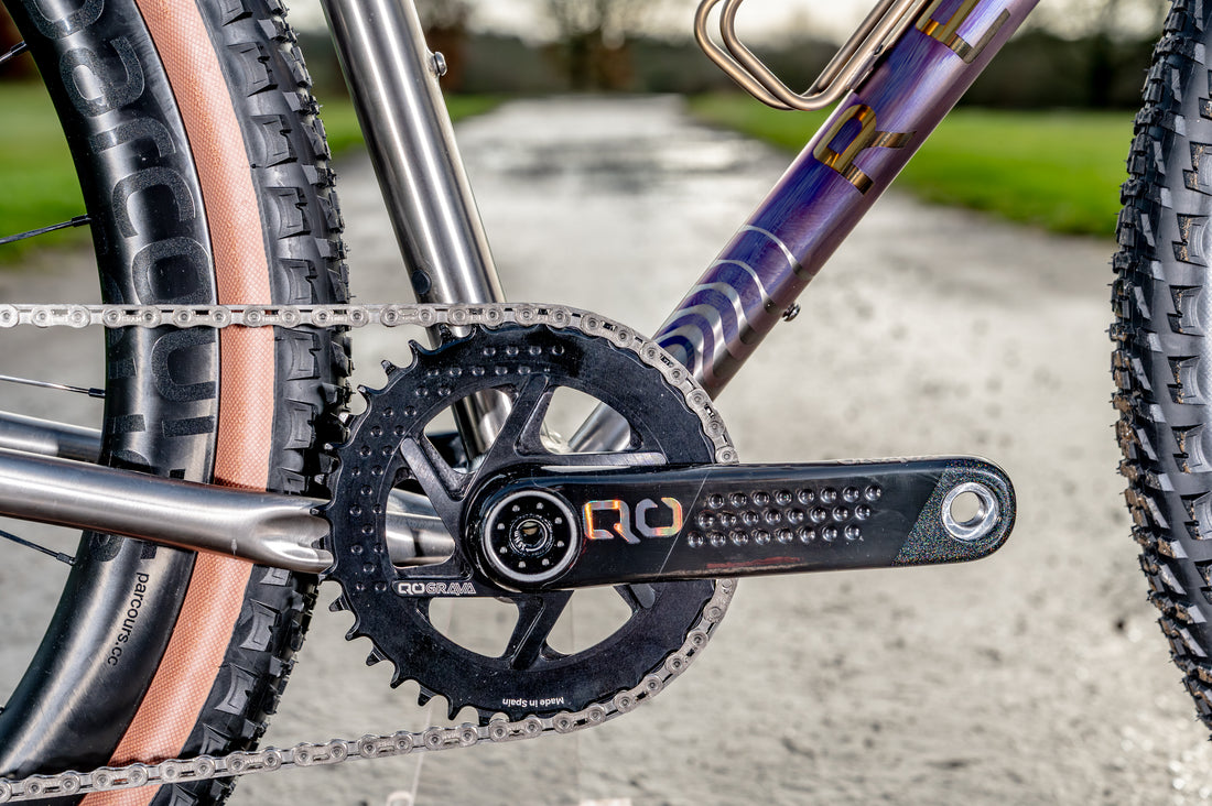 Close-up of a bicycle crankset with a black chainring and black crank arm featuring gold 'RQ' branding, attached to a purple and gold frame. The bike is outdoors on a paved path with tires visible on both sides.