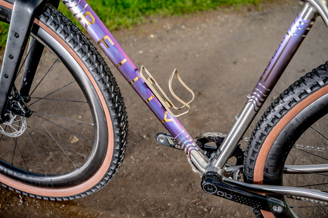 Close-up of a bicycle frame and wheels on a dirt path. The frame has a purple and gold gradient finish with stylized lettering, and the tires have pink sidewalls.