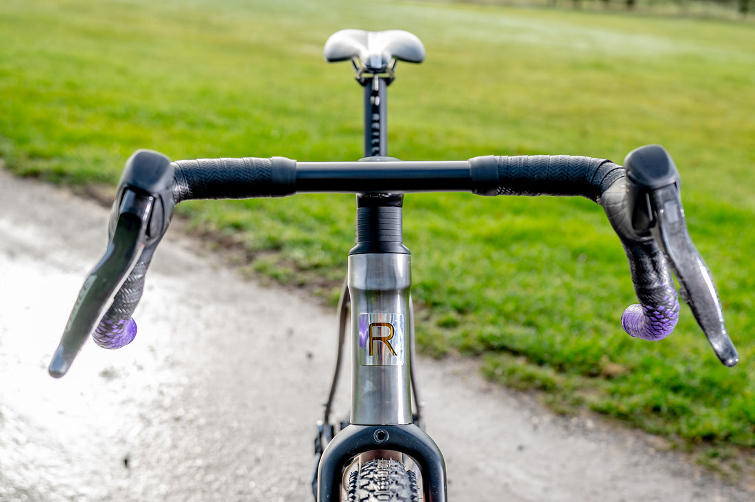 Close-up of a bicycle drop handlebar setup on a gravel path with a blurred green grass background. The handlebar tape and brake levers show a distinctive purple and black pattern, and a silver frame with a gold 'R' logo is visible.