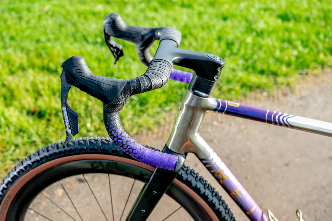 Close-up of a gravel bicycle handlebar area with black drop handlebars, purple tape wrapping, a silver and purple frame, and a blurred green grass background.