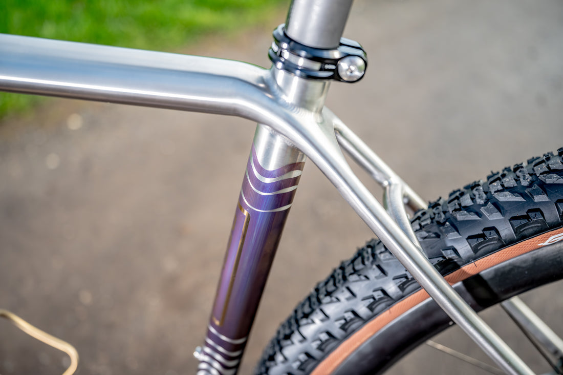 Close-up detail of a bicycle frame with a metallic silver tubular structure and a purple down tube featuring white stripes, with part of a black knobby tire visible.