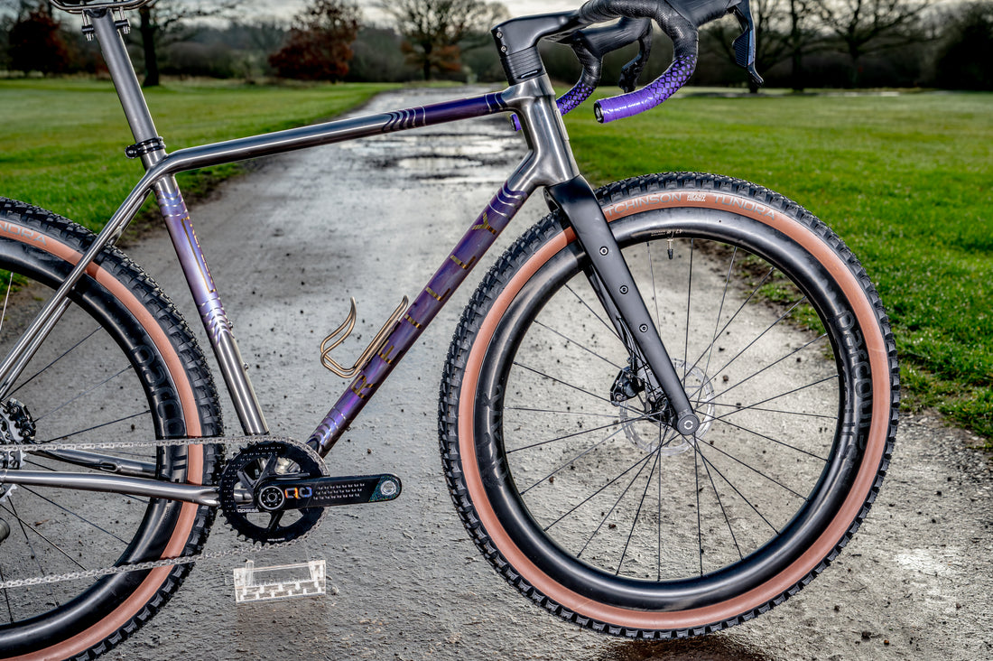 A close-up of a gravel bicycle with a metallic purple and gray frame, black handlebars, and tan sidewall tires, positioned on a wet paved path with a grassy park in the background.