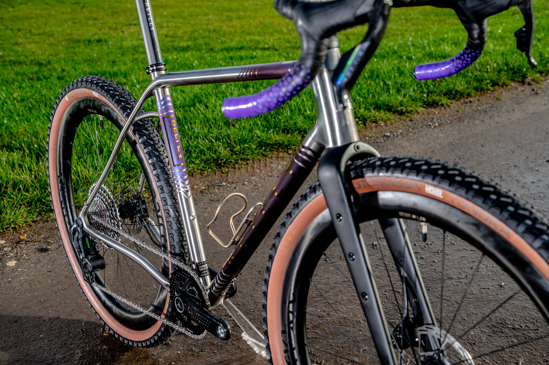 Close-up of a gravel bicycle with a metallic silver frame, black components, and purple handlebar tape, parked on a wet paved path with grass in the background.
