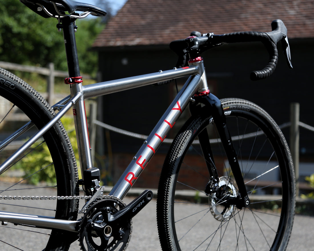 Close-up of a silver Perilly gravel bike frame with red lettering, black handlebars, and black tires, photographed outdoors with a blurred background of trees and a building.