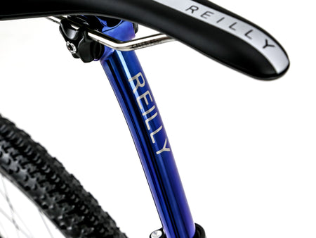 Close-up of a Reilly bicycle frame in metallic blue with silver lettering, attached to a black saddle with white branding, on a white background.