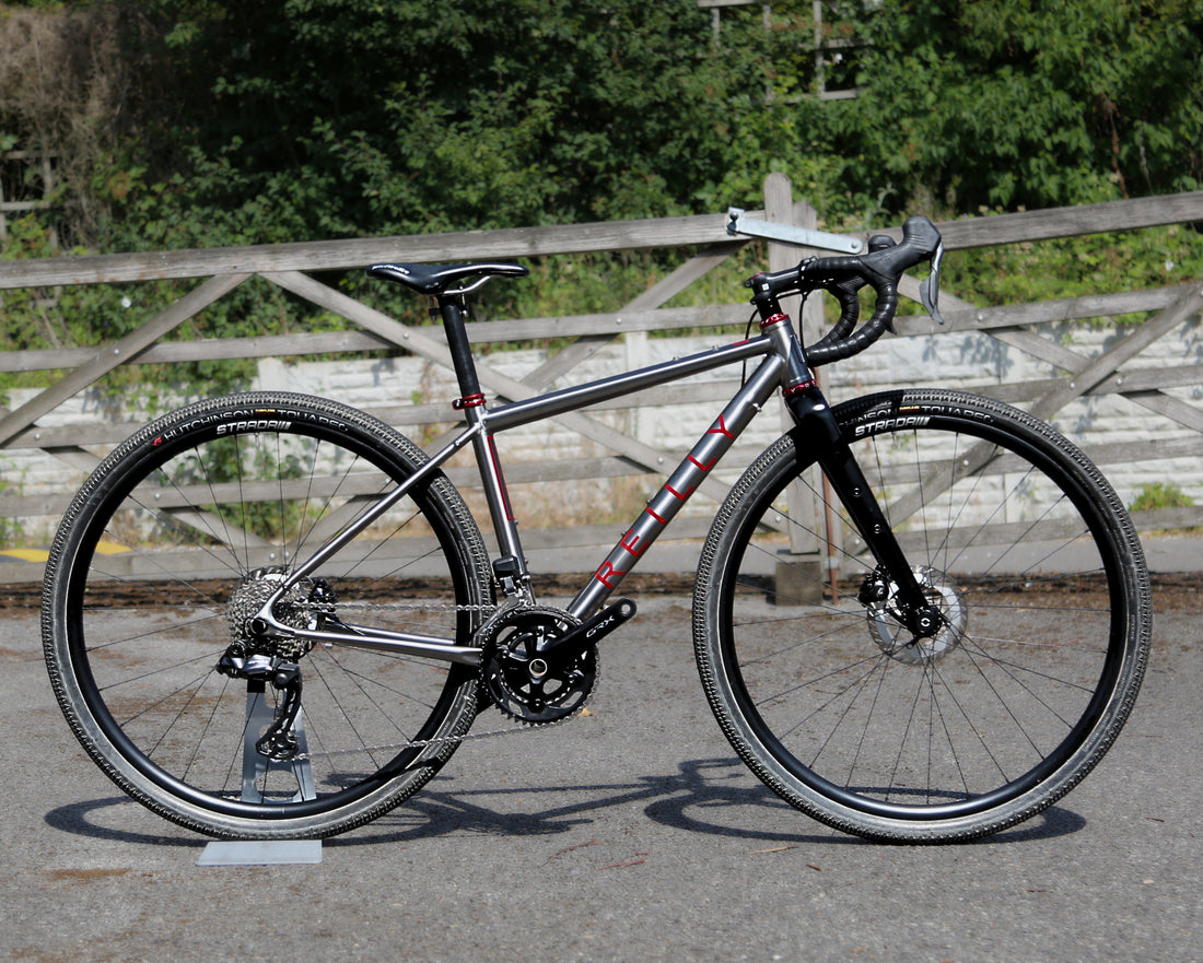 A drop-bar road bicycle with a matte dark gray/silver frame and black components, photographed outdoors on a paved surface with a wooden fence and greenery in the background.