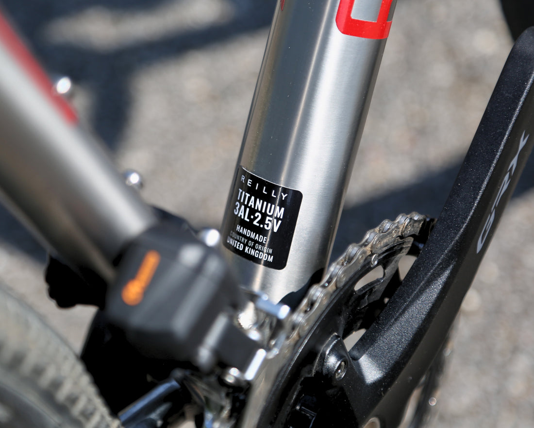Close-up of a bicycle frame component showing a silver Neill Titanium 3Ax 2.5V rear shock absorber with a black label, alongside a black crank arm and chainring in an outdoor setting.