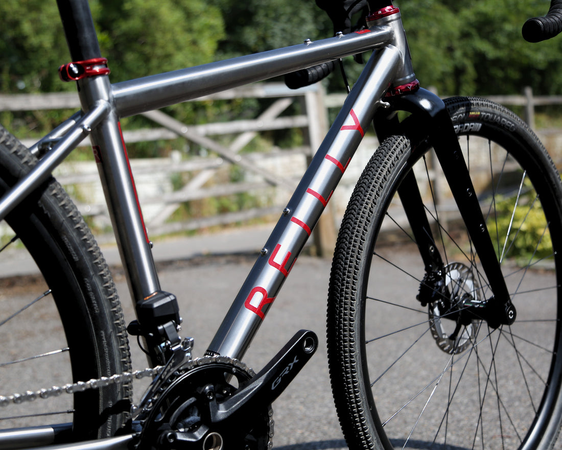 Close-up of a gravel bicycle frame in metallic silver with red accents and the brand name 'REILLY' printed on the down tube, set outdoors on a paved path with blurred greenery and fencing in the background.