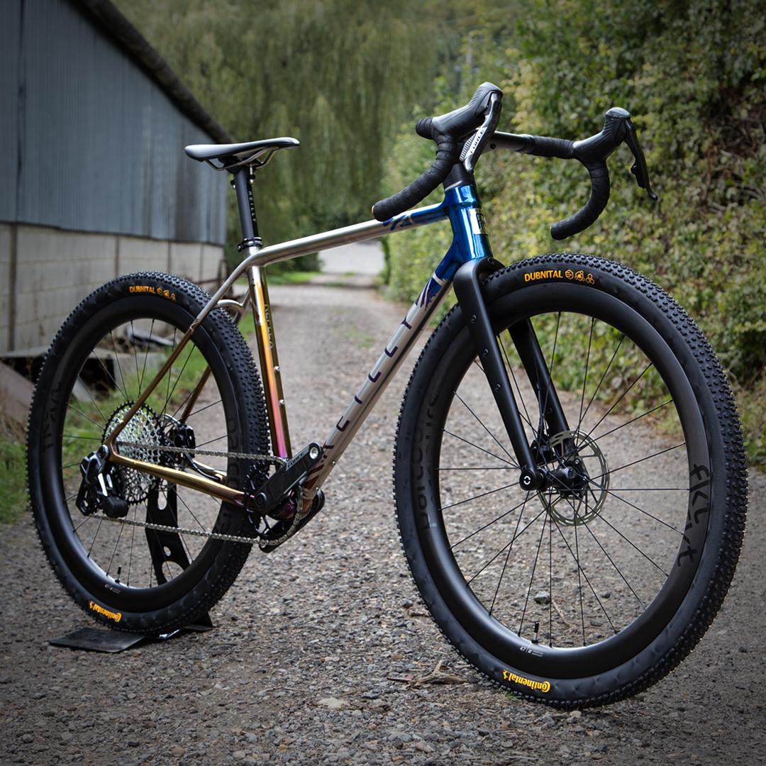 A high-end gravel bicycle with a metallic silver and blue frame, black components, and knobby tires, photographed outdoors on a gravel path next to a building.
