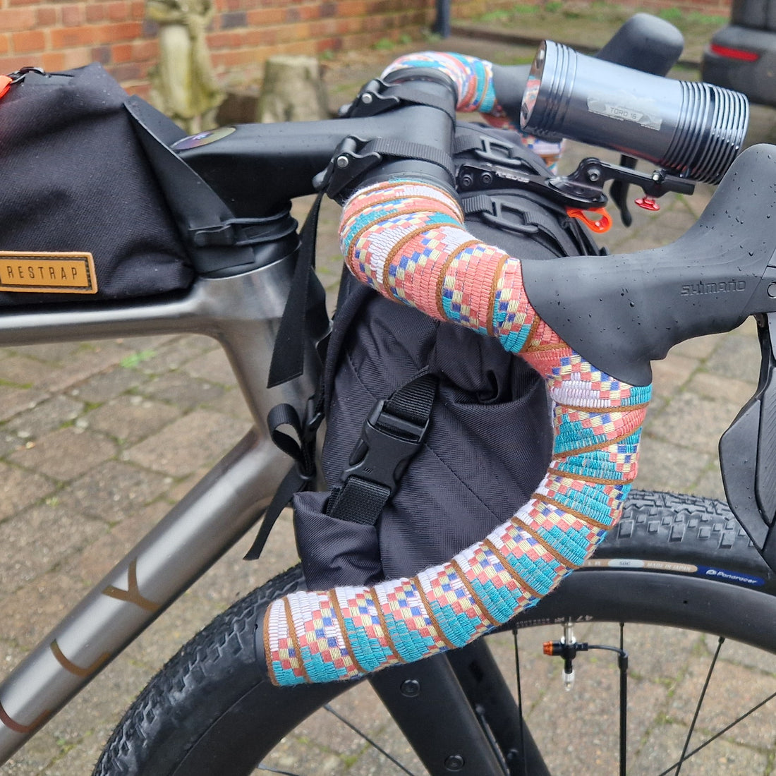 Bicycle handlebar with colorful grips and black bag on a brick-paved street.