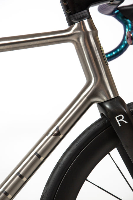 Close-up of a bicycle frame in a matte metallic gray finish, showing the junction with the fork and part of the black wheel against a white background.