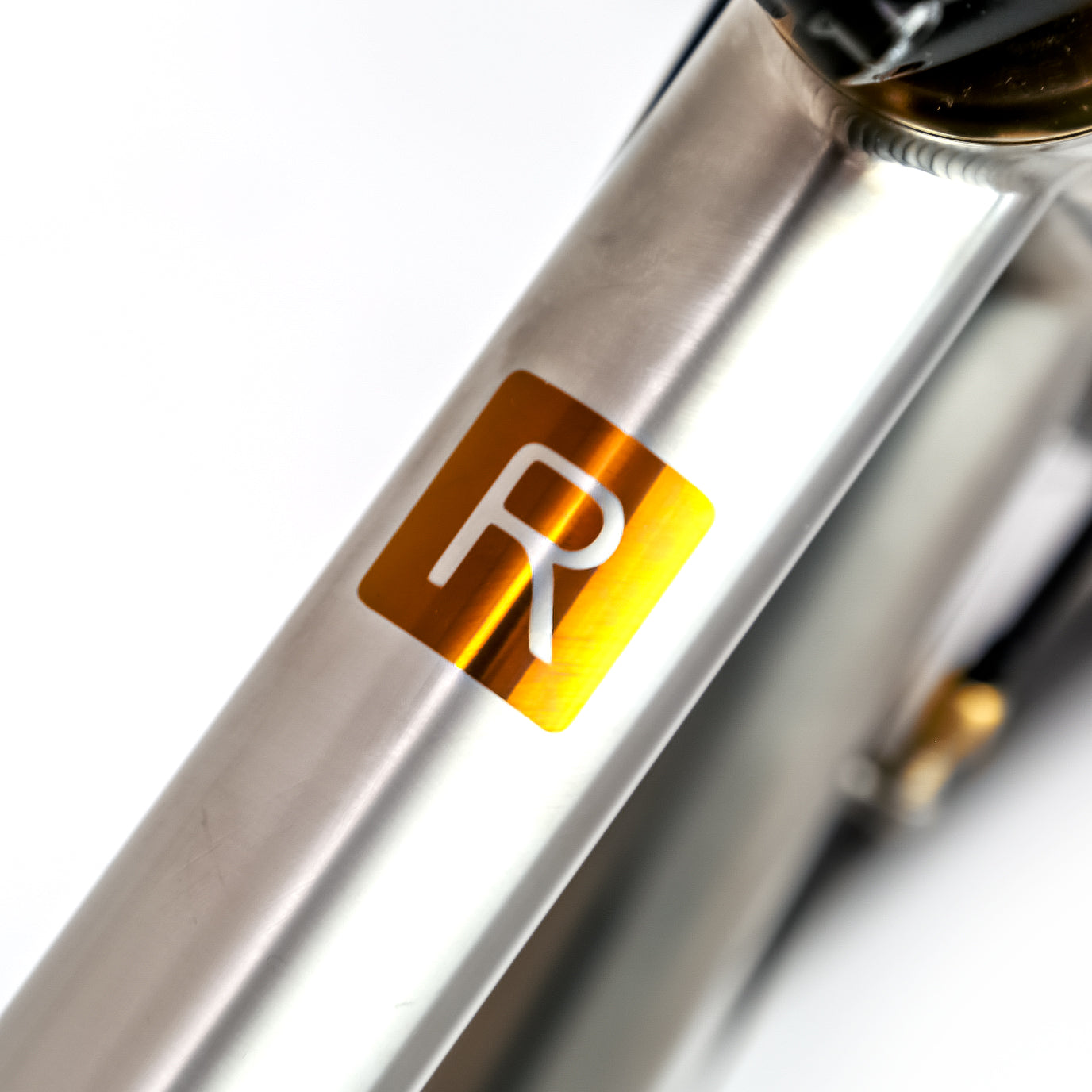 Close-up of a metallic bicycle frame with a square gold decal featuring a white stylized letter R, on a white background.