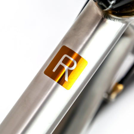 Close-up of a metallic bicycle frame with a square gold decal featuring a white stylized letter R, on a white background.
