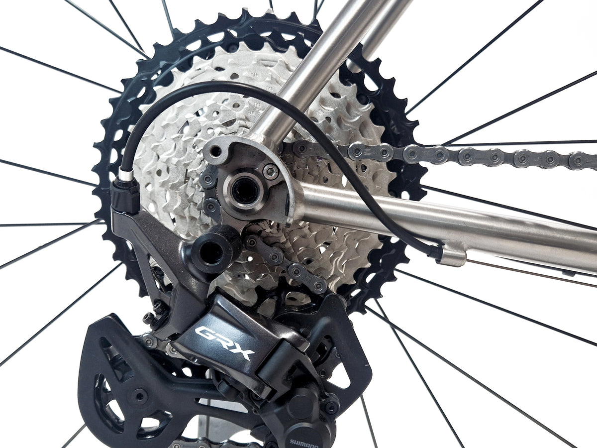 Close-up of a bicycle rear derailleur and cassette with a silver chainring, black chain, and black GRX derailleur on a white background.
