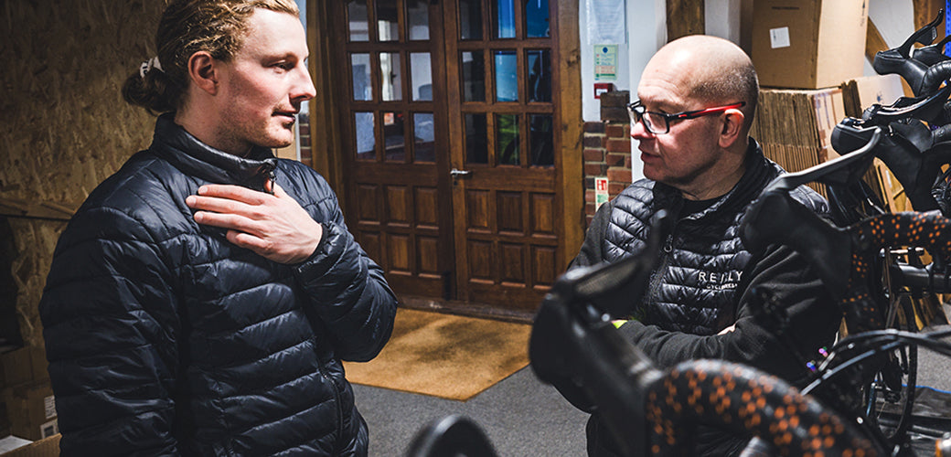 Two men in black puffer jackets standing and talking indoors near a bicycle in a shop or workshop setting.