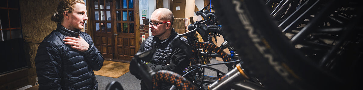 Two men in black puffer jackets standing and talking indoors near a bicycle in a shop or workshop setting.