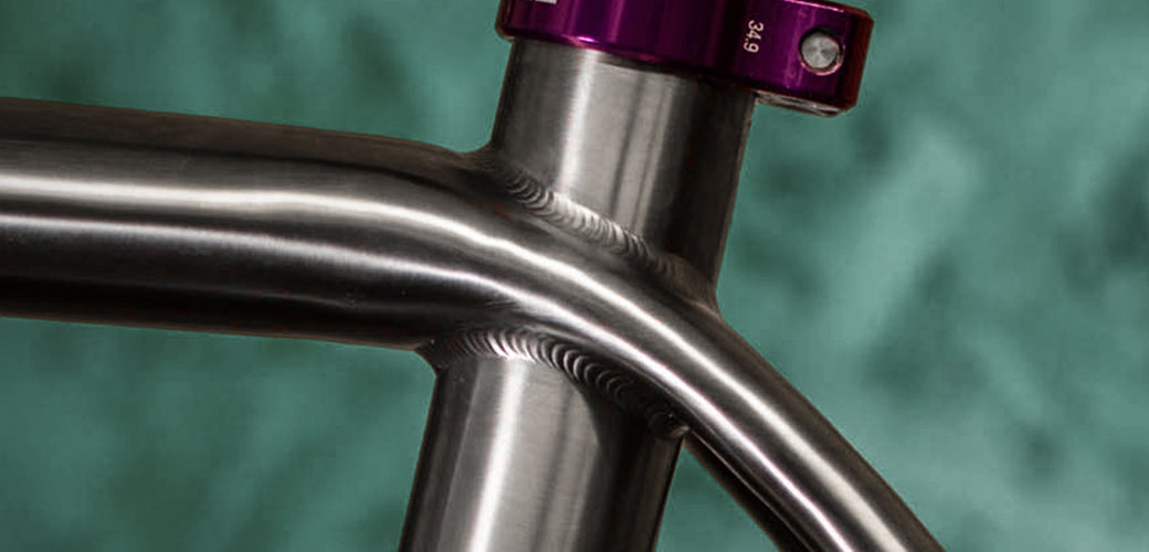 Close-up of a metallic bicycle frame showing the seat tube junction and a purple headset, set against a textured teal background.