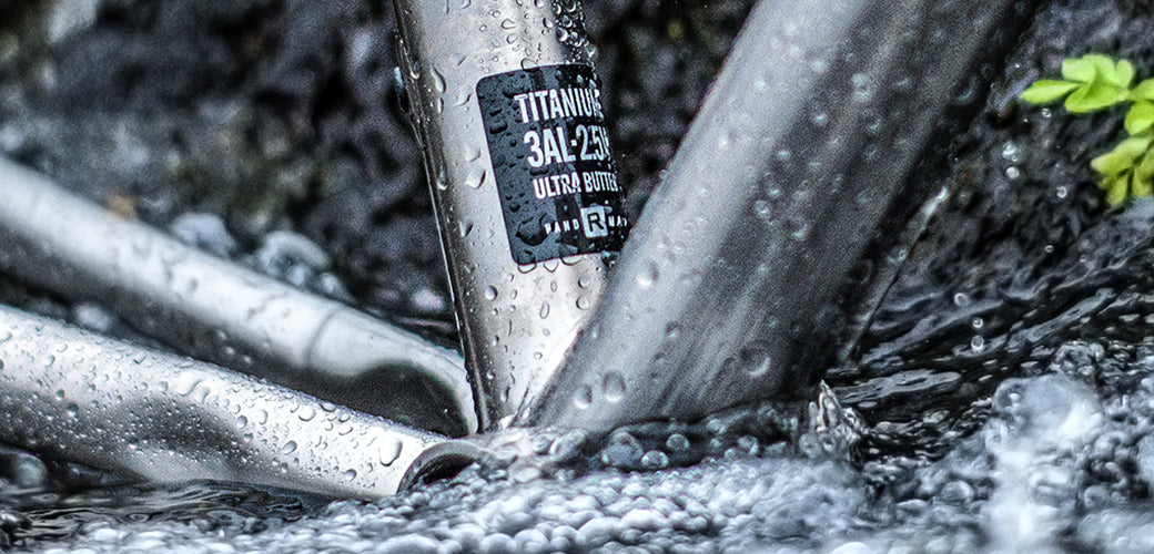 Close-up of a metallic bicycle frame in water, with a black label reading 'TITANIUM 3AL-ZR ULTRA BOTTLE' visible on the seat tube.