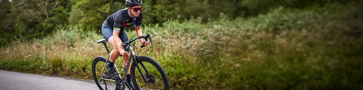 A cyclist in a black and green jersey rides a road bike on a paved path with tall green grass and trees in the background.