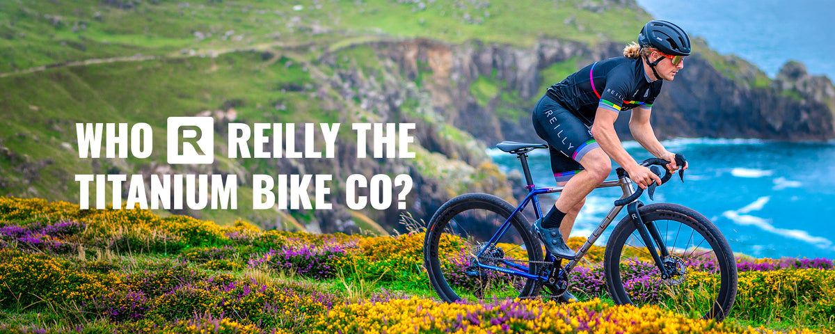 A cyclist wearing a black helmet and black cycling kit rides a dark blue titanium road bike along a grassy coastal path with purple and yellow wildflowers and blue ocean in the background. Large white text on the left reads 'WHO REILLY THE TITANIUM BIKE CO?'.