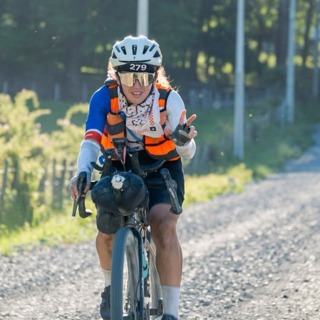 A female cyclist makes the peace sign while cycling on gravel.
