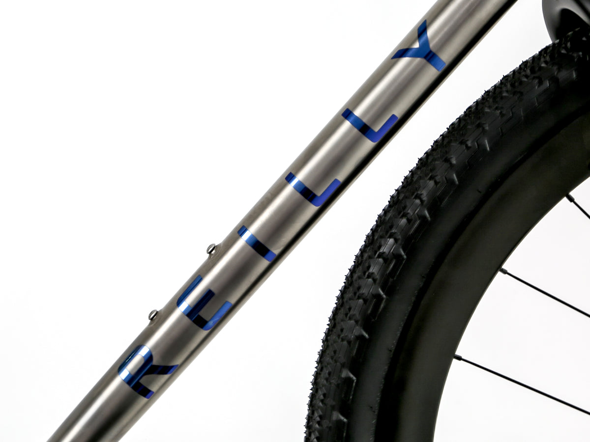 Close-up of a bicycle frame tube with the brand name 'KILLY' in blue on a metallic silver finish, alongside a black tire and wheel against a white background.