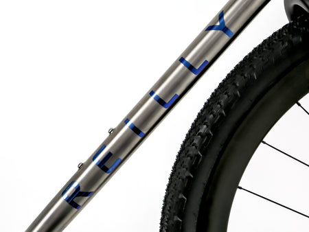 Close-up of a bicycle frame tube with the brand name 'KILLY' in blue on a metallic silver finish, alongside a black tire and wheel against a white background.