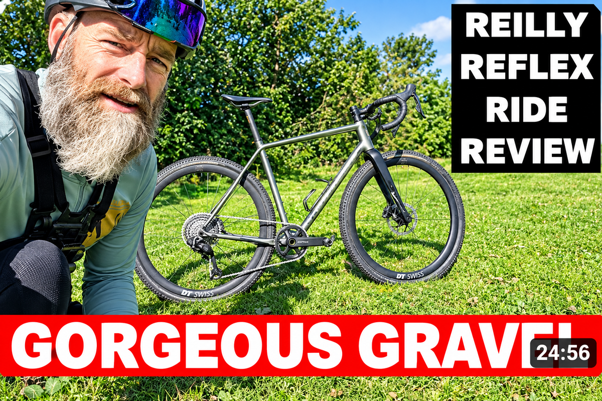 Load video: A man with a gray beard and helmet lies on grass next to a gray gravel bicycle, with large text overlays reading 'REILLY REFLEX RIDE REVIEW' and 'GORGEOUS GRAVEL'.