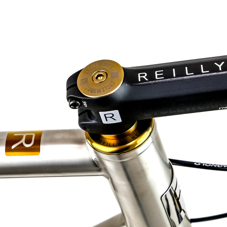 Close-up of a bicycle headset with a black Reilly stem, gold headset cup and collar, and a silver frame post on a white background.
