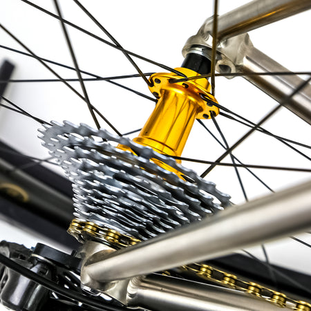 Close-up of a bicycle's rear wheel hub and cassette, featuring a shiny gold anodized hub, silver metal cassette cogs, a gold chain, and silver spokes against a white background.