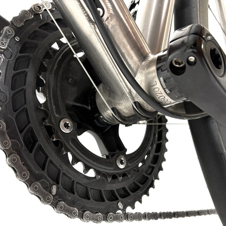 Close-up of a bicycle crankset and chainring, showing a black chainring with a silver chain, part of a silver frame, and a black bottom bracket on a white background.