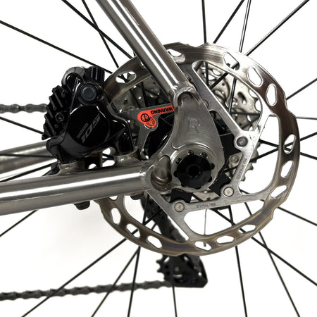 Close-up of a bicycle's rear disc brake assembly, featuring a silver metallic rotor, black brake caliper and Reilly branding.