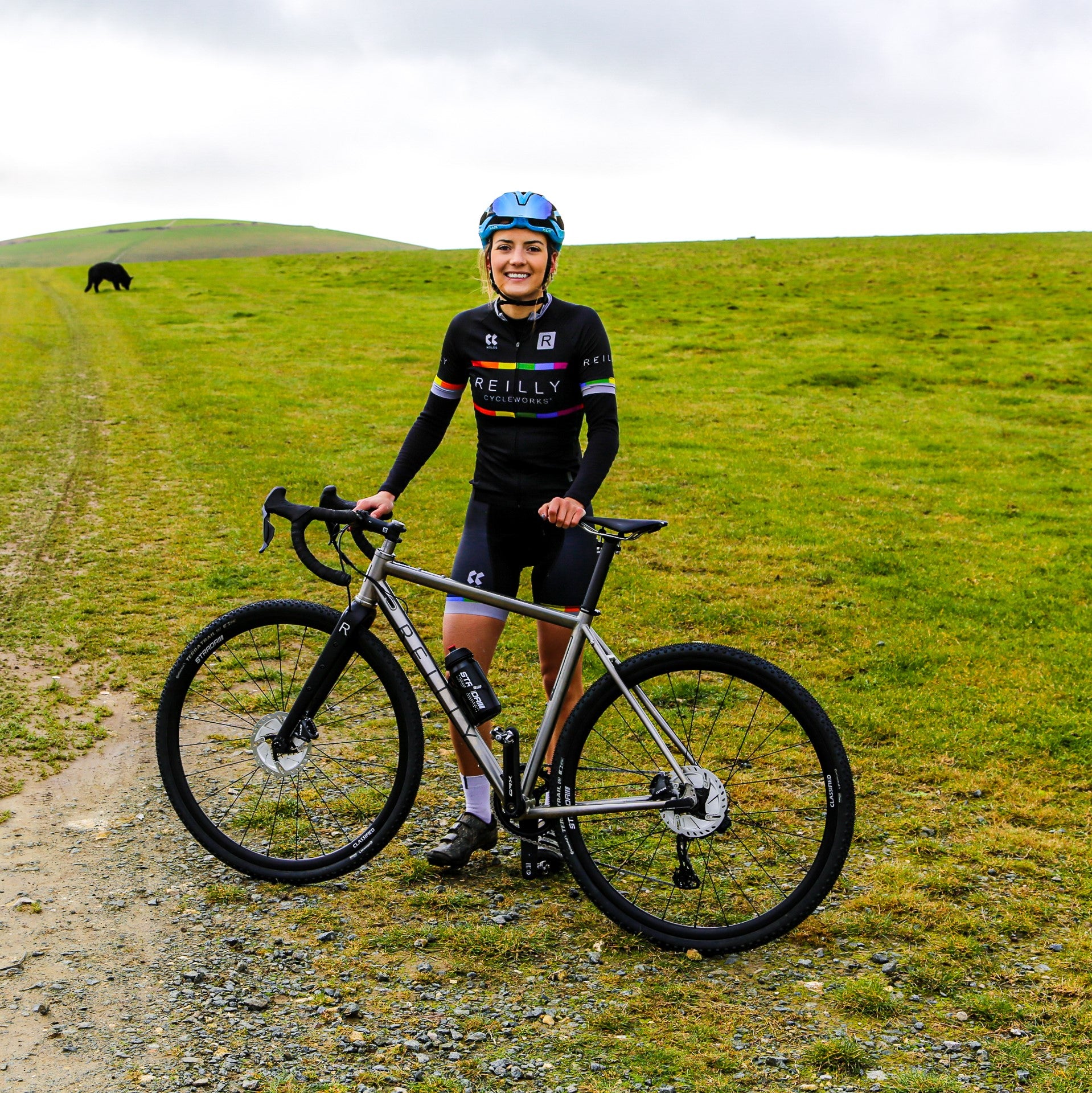 Photo of a blonde lady standing proudly next to her Reilly Bike in a green feild.